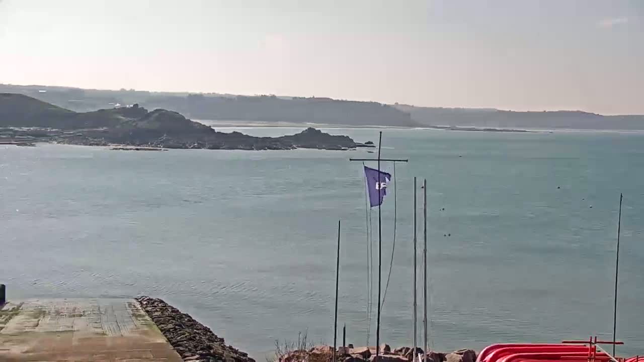 A calm, sunny day overlooks a body of water with a rocky coastline and a flagpole with a purple flag.