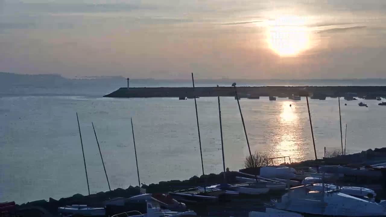 A calm body of water with numerous boats at a harbor reflects the setting sun under a mostly clear sky.