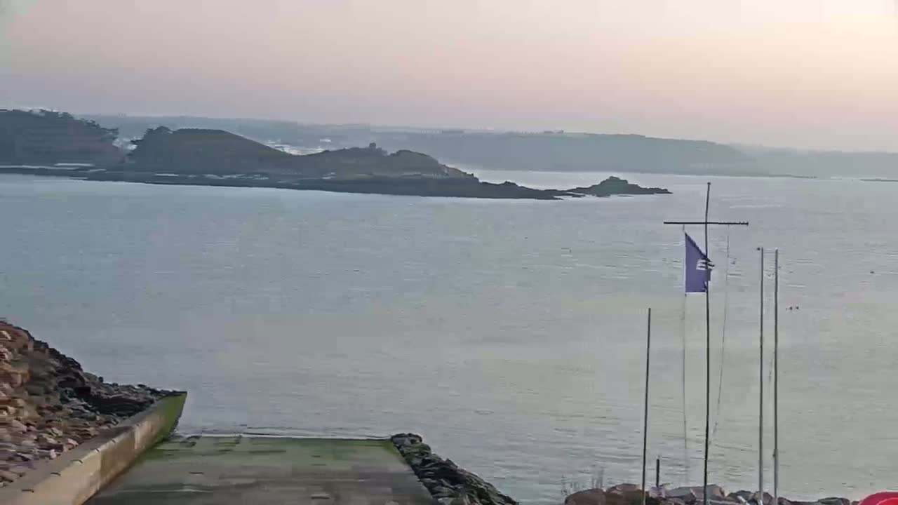 A calm, hazy body of water is seen from a high vantage point, featuring a small island in the distance and a boat ramp in the foreground with flagpoles.