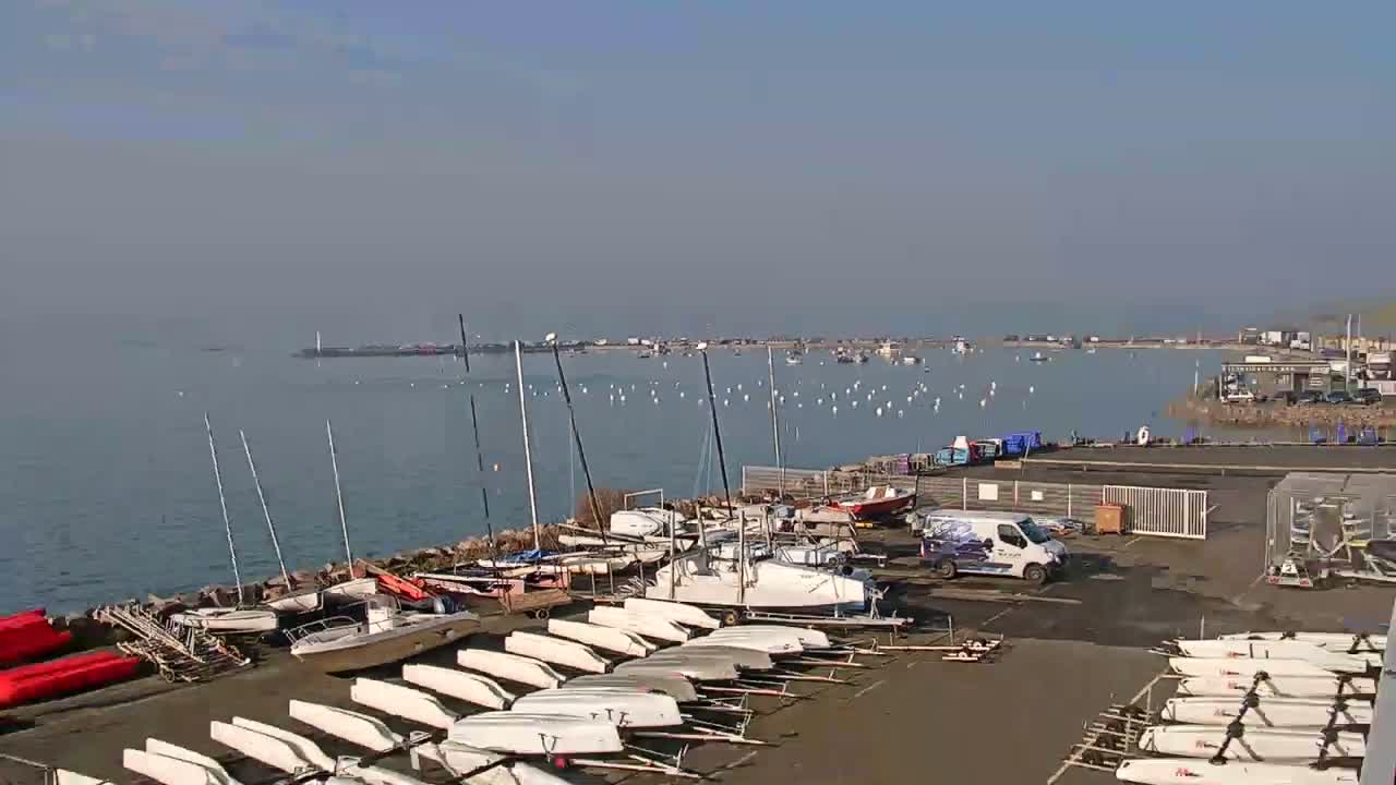 Numerous sailboats and small motorboats are docked at a harbor on a hazy, sunny day, with additional boats visible in the water beyond.