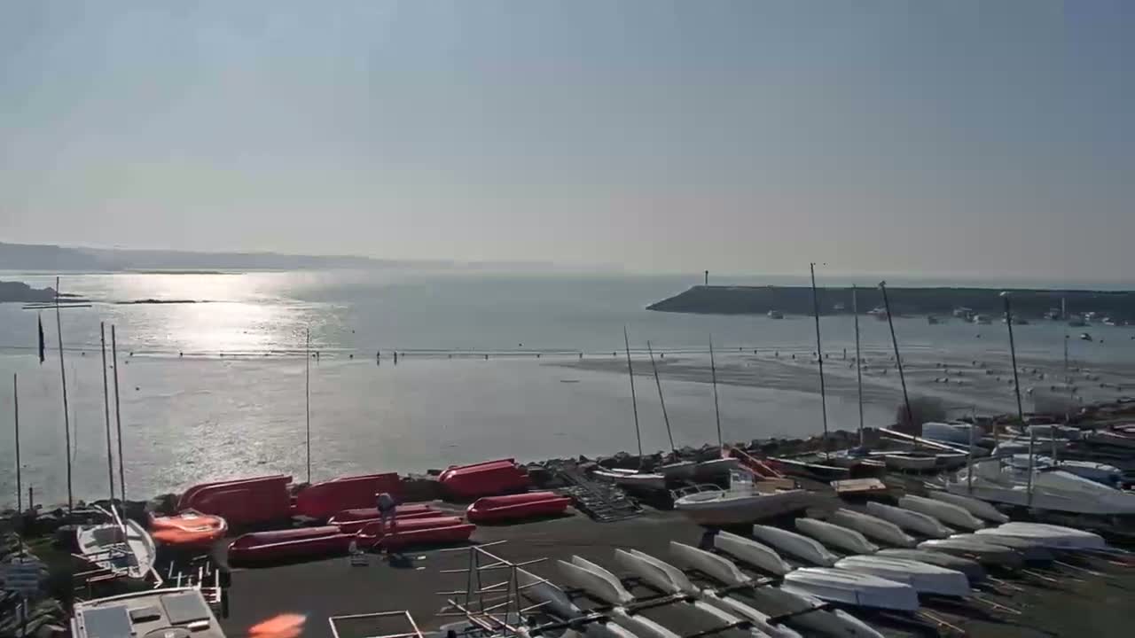 A sunny day reveals a calm bay with numerous sailboats and small crafts docked along a rocky shoreline, many more boats are visible in the distance.