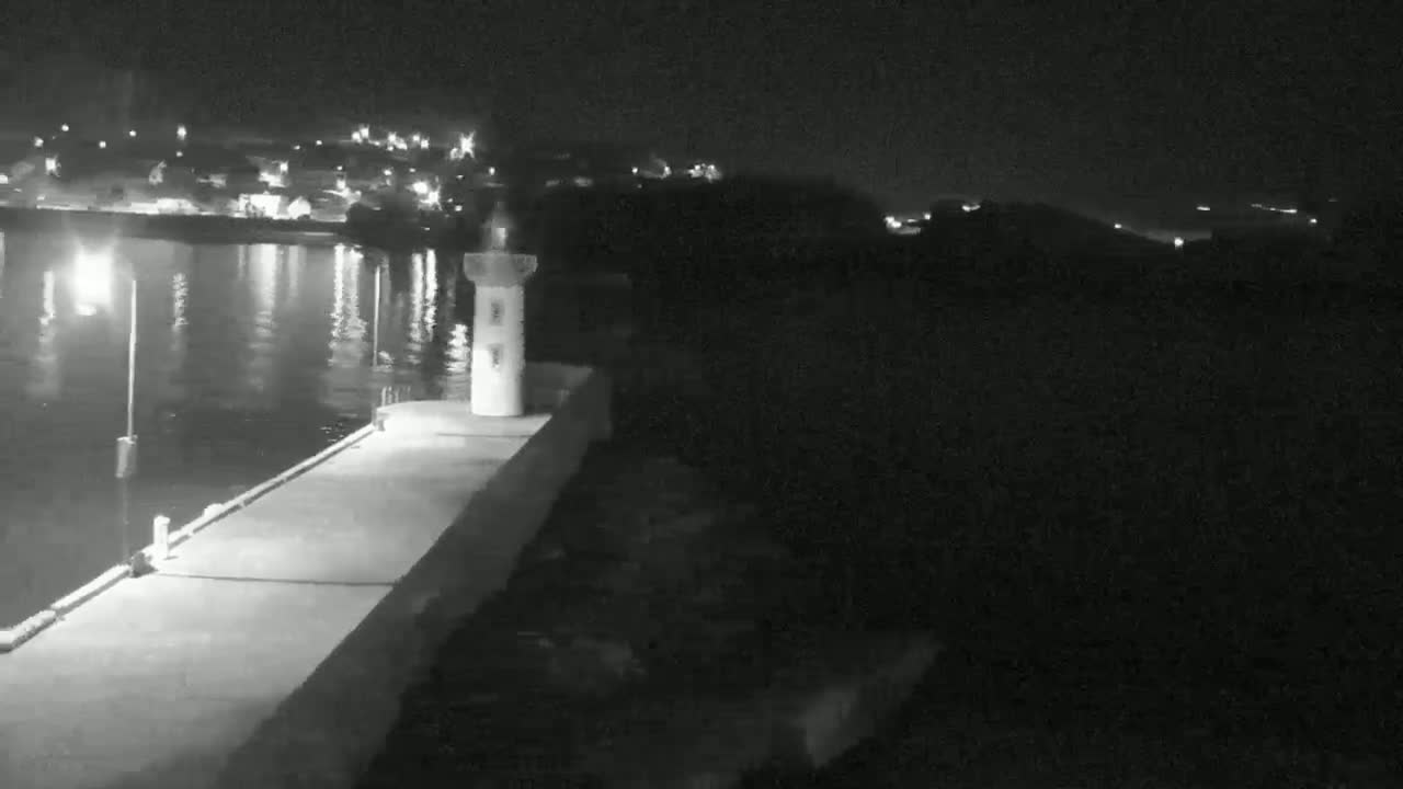 A nighttime view shows a concrete pier extending into calm water, with a small lighthouse visible on the pier and a town's lights faintly illuminating the background.