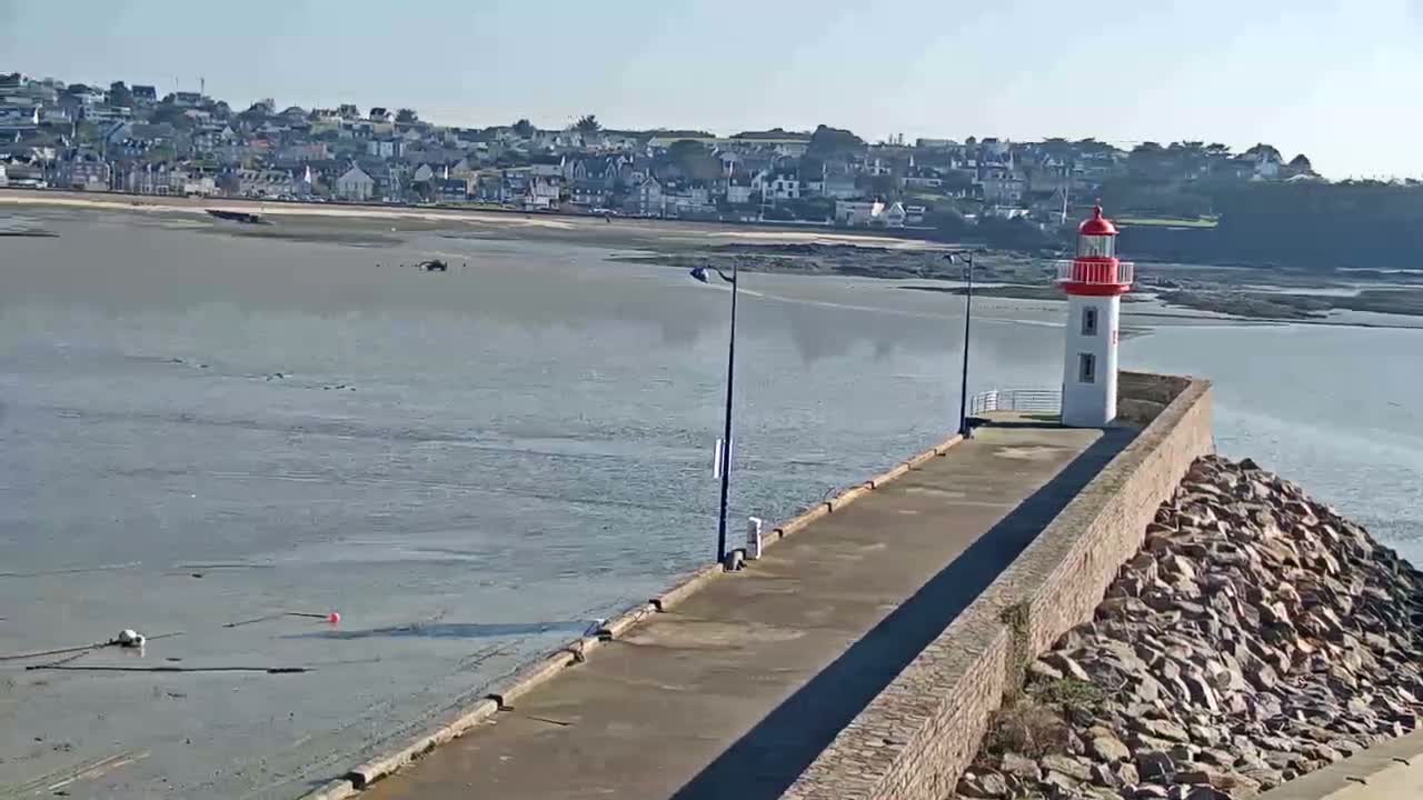 A long stone pier extends from a rocky shoreline toward a small lighthouse under a clear, sunny sky, with a town visible in the distance and calm water in the foreground.