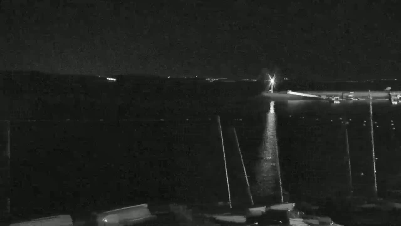 A nighttime, low-light view of a body of water with several masts of boats visible in the foreground and a light source across the water.
