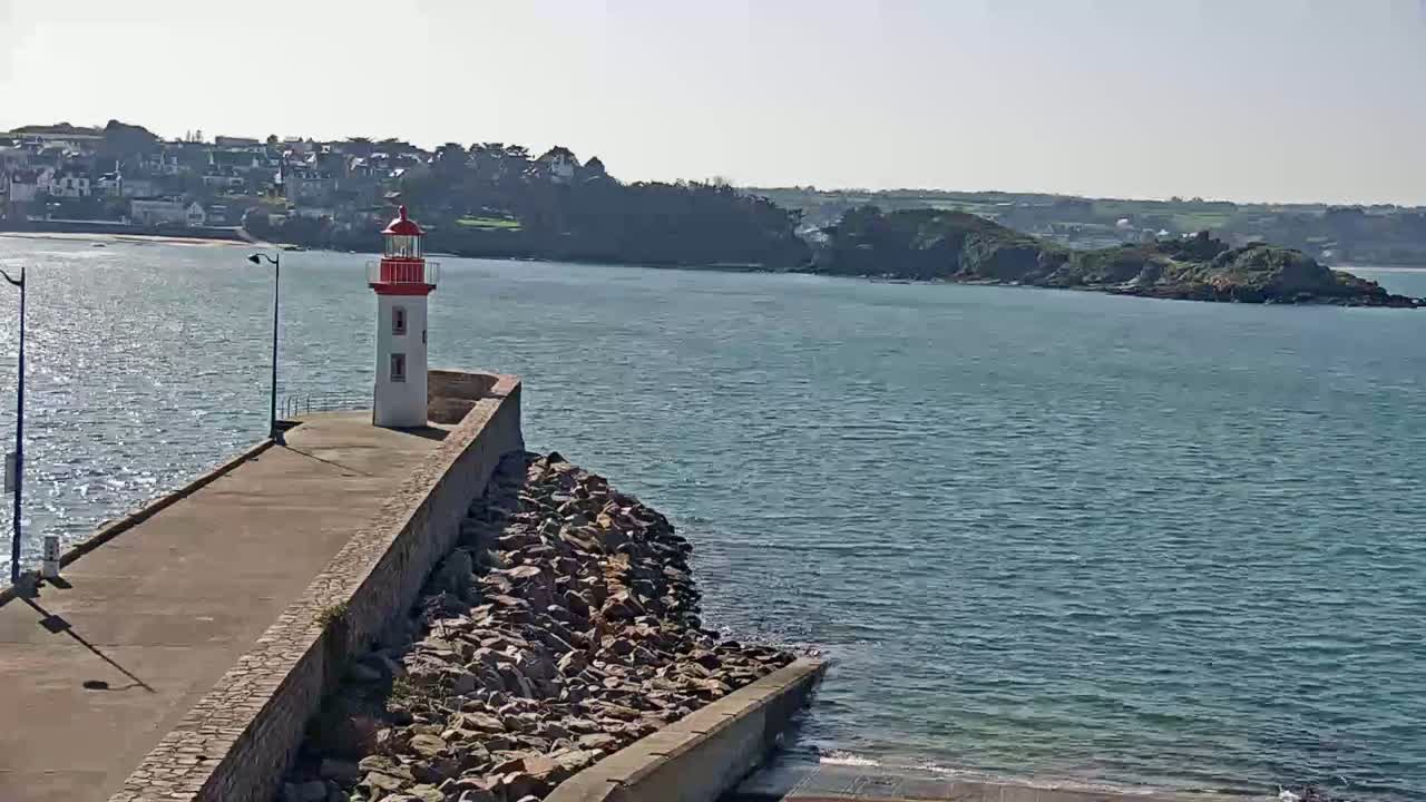 A red and white lighthouse stands at the end of a stone seawall overlooking calm blue water and a distant coastline on a sunny day.