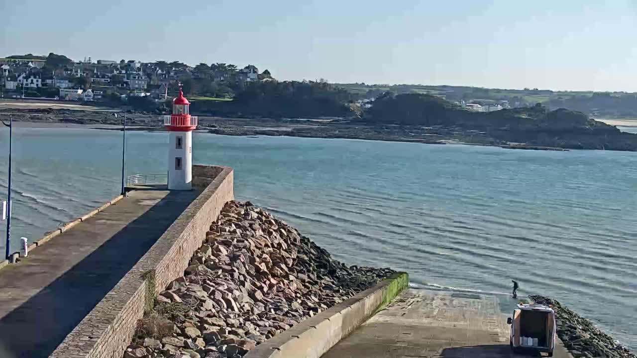 A red and white lighthouse stands on a stone pier overlooking a calm, blue-green sea on a sunny day.