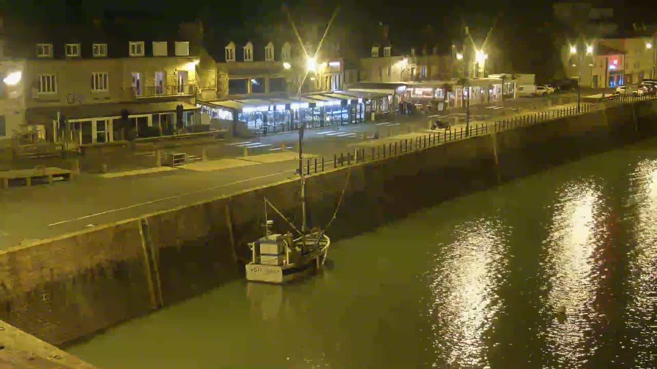 A small fishing boat is moored in a calm harbor at night, alongside a waterfront lined with buildings and streetlights reflecting in the water.