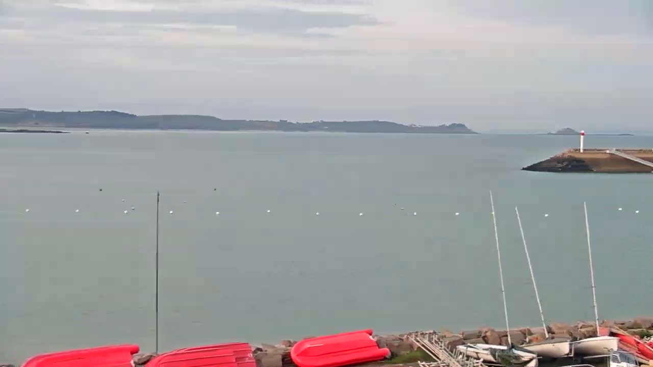 A calm, overcast day reveals a body of water with a distant shoreline, a lighthouse on a small island, and several small boats moored near a rocky shore.