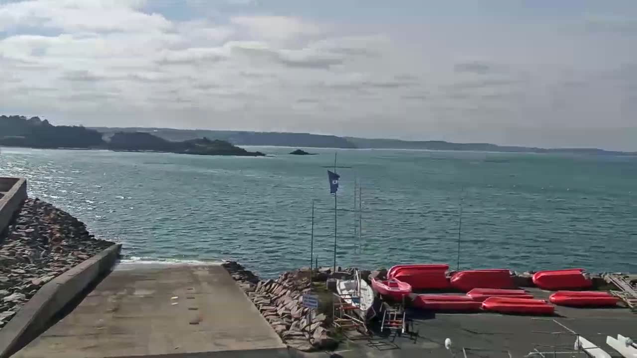 A boat ramp leads to a calm, blue body of water under a partly cloudy sky, with several red small boats resting on land nearby.