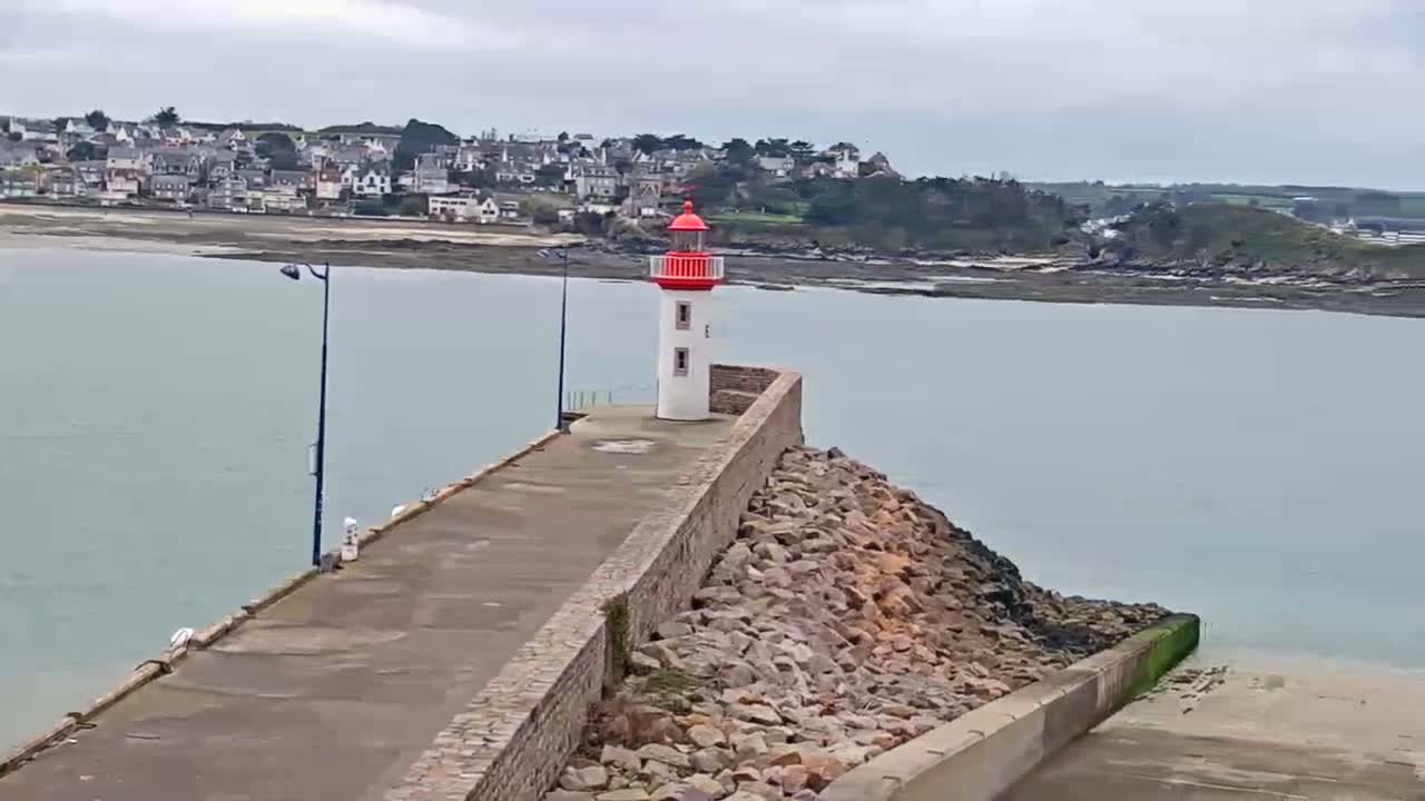 A white and red lighthouse stands at the end of a stone jetty extending into a calm, light-grey body of water, with a town visible in the background on an overcast day.