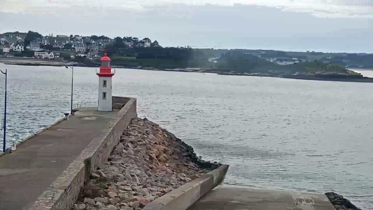 A white and red lighthouse stands on a rocky breakwater overlooking a calm body of water, with a town visible in the distance under an overcast sky.