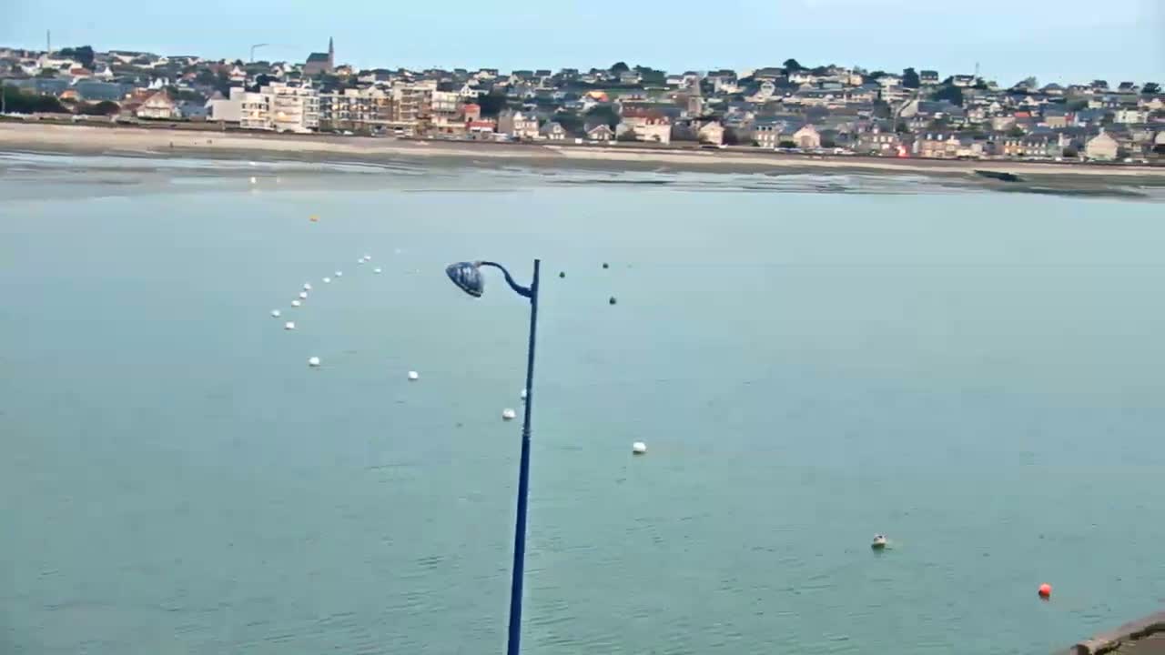 A coastal town is visible in the distance across calm, light-blue water dotted with white buoys, a single lamp post stands in the foreground.
