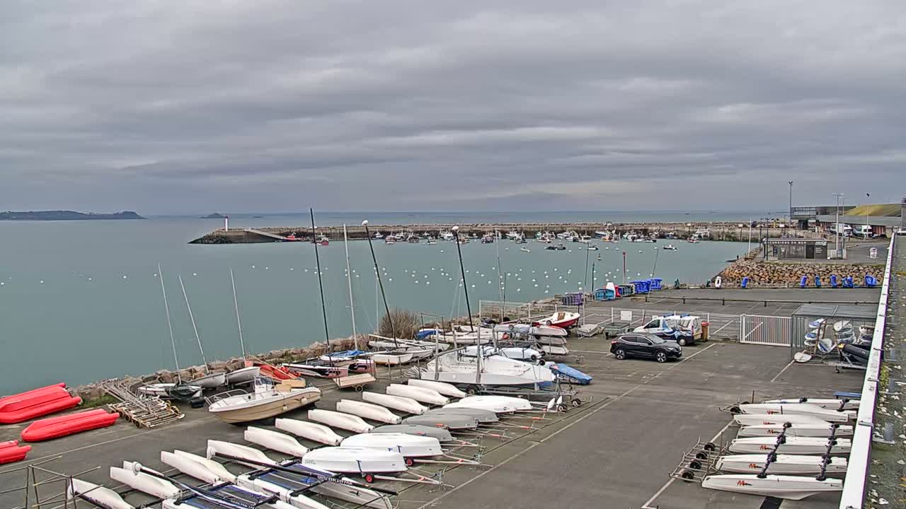 A harbor filled with numerous sailboats and motorboats under a cloudy sky, with additional boats and kayaks stored on land nearby.
