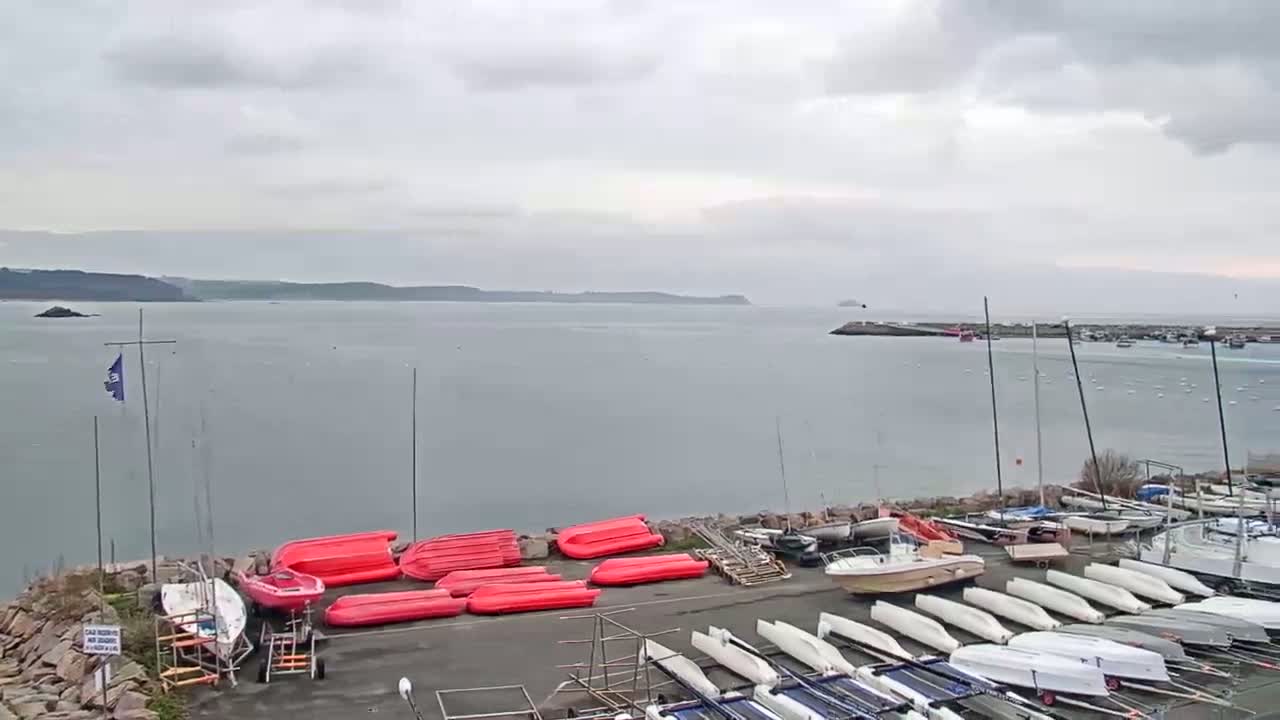 A harbor filled with numerous sailboats and small powerboats under a cloudy sky, with a calm body of water extending to a distant shoreline.
