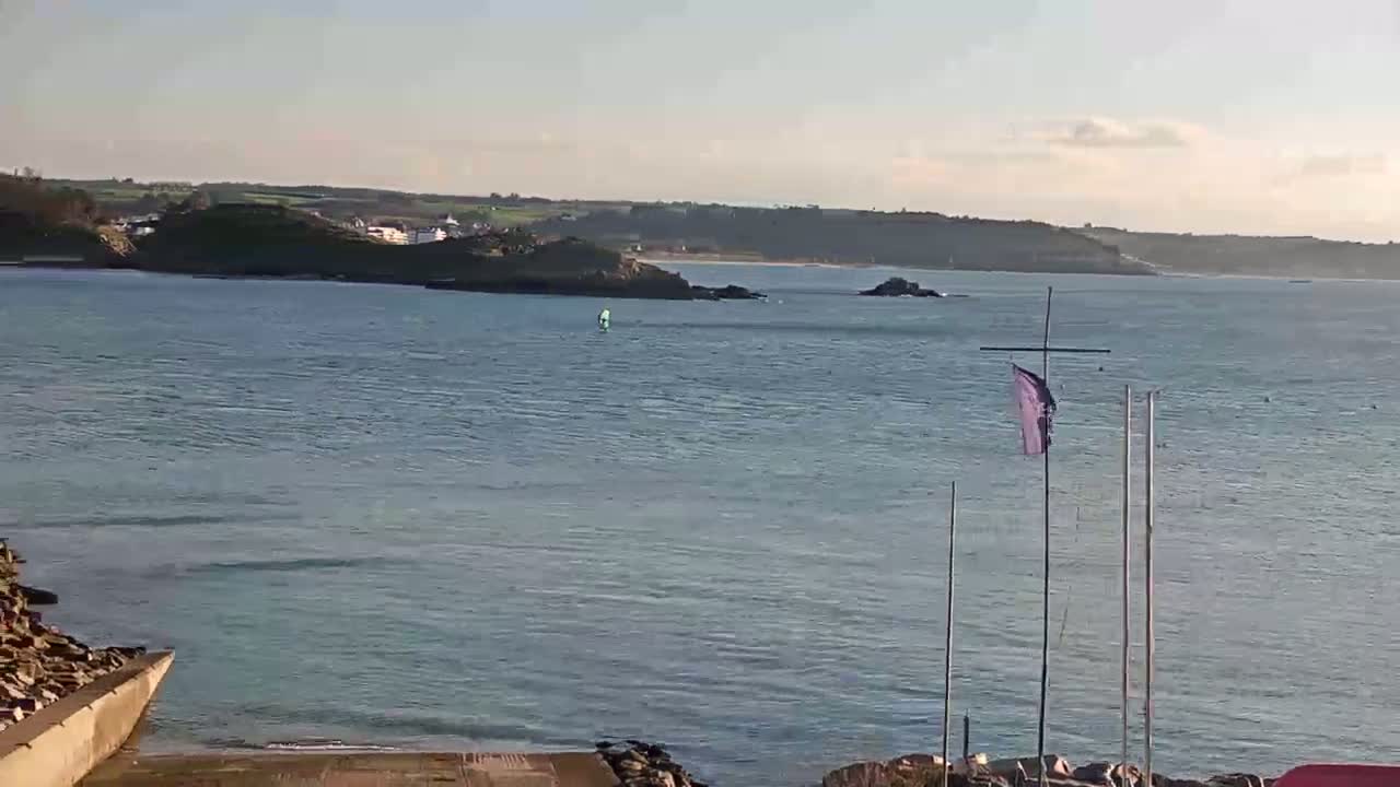 A calm body of water stretches out to a rocky shoreline under a clear, sunny sky, with a small sailboat visible in the distance.
