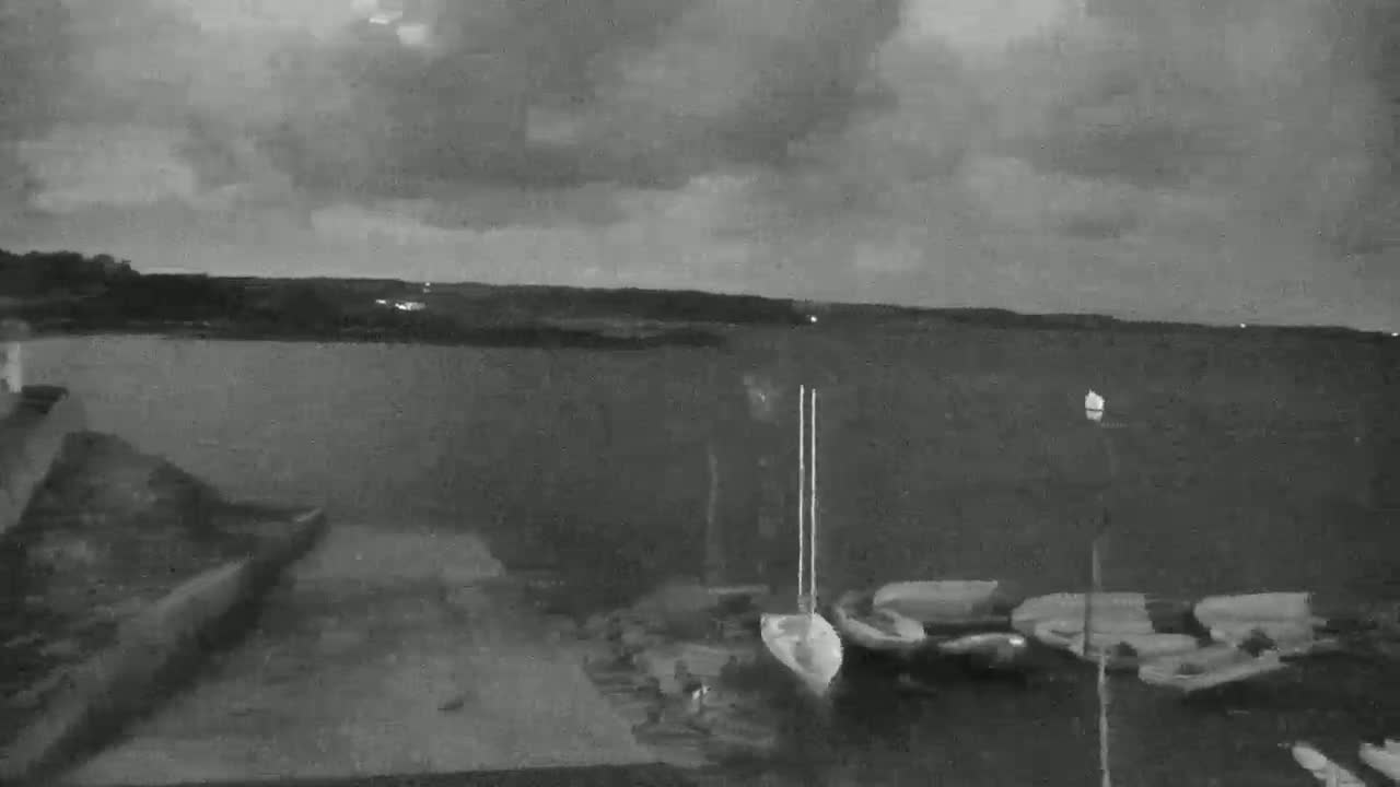 A grayscale image shows a calm body of water under a cloudy, dark sky at night, with several small boats moored near a shoreline.