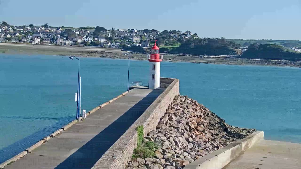 A small, red and white lighthouse stands on a rocky breakwater extending from a coastal town on a sunny day with calm, turquoise water.