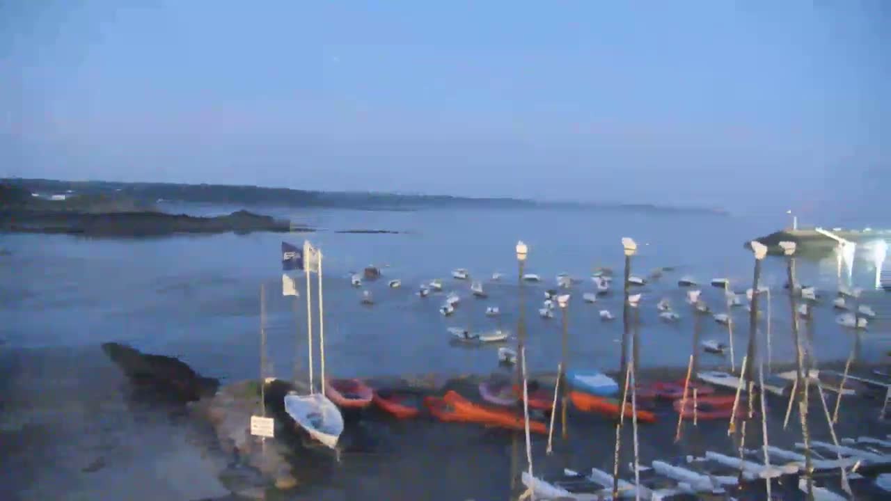 Many small sailboats and other watercraft are moored in a calm bay under a twilight sky.