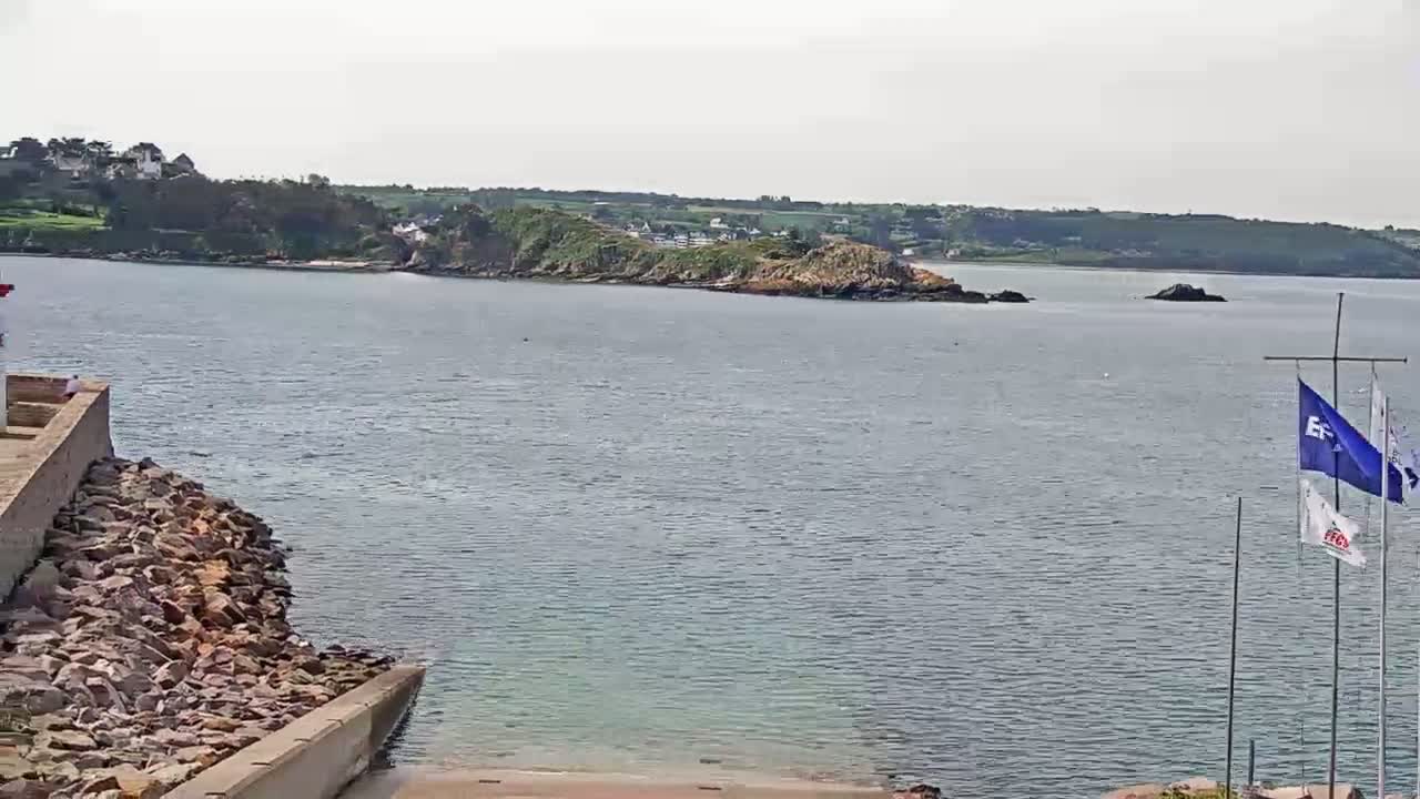A rocky shoreline with a calm body of water stretches toward a small, verdant island under a cloudy sky.