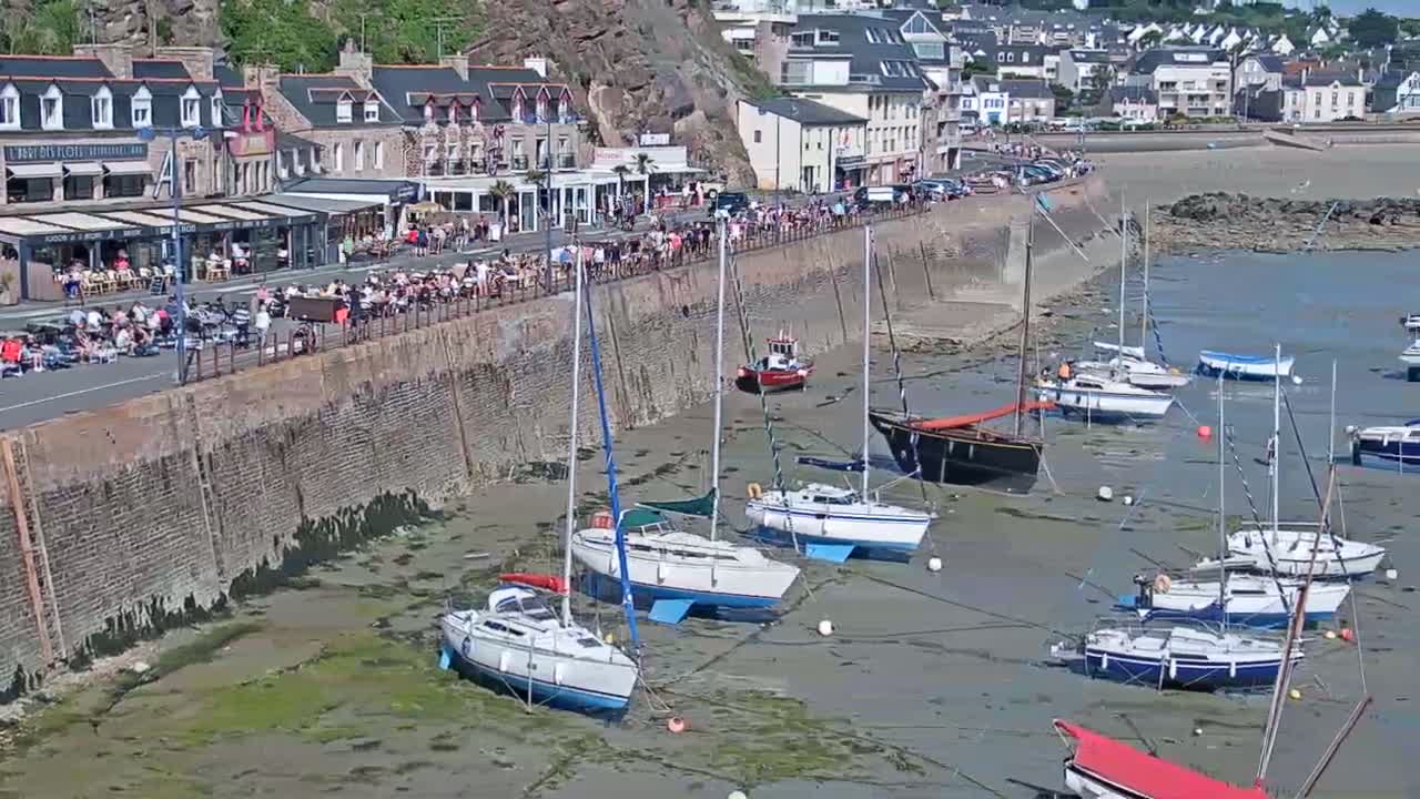 A seaside town with a stone wall along a harbor where many sailboats are sitting in shallow, muddy water at low tide, under a sunny sky.