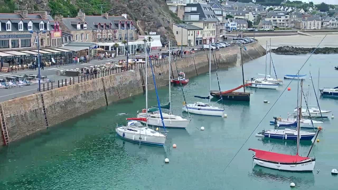 Several sailboats are moored in a calm, teal harbor next to a stone seawall lined with restaurants and buildings.