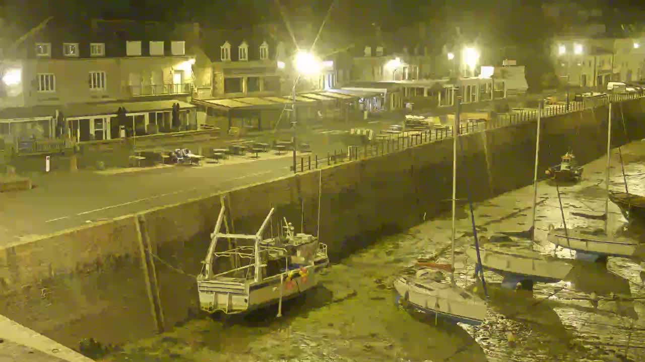 At night, several boats are docked in shallow water next to a stone wall and buildings with outdoor seating.