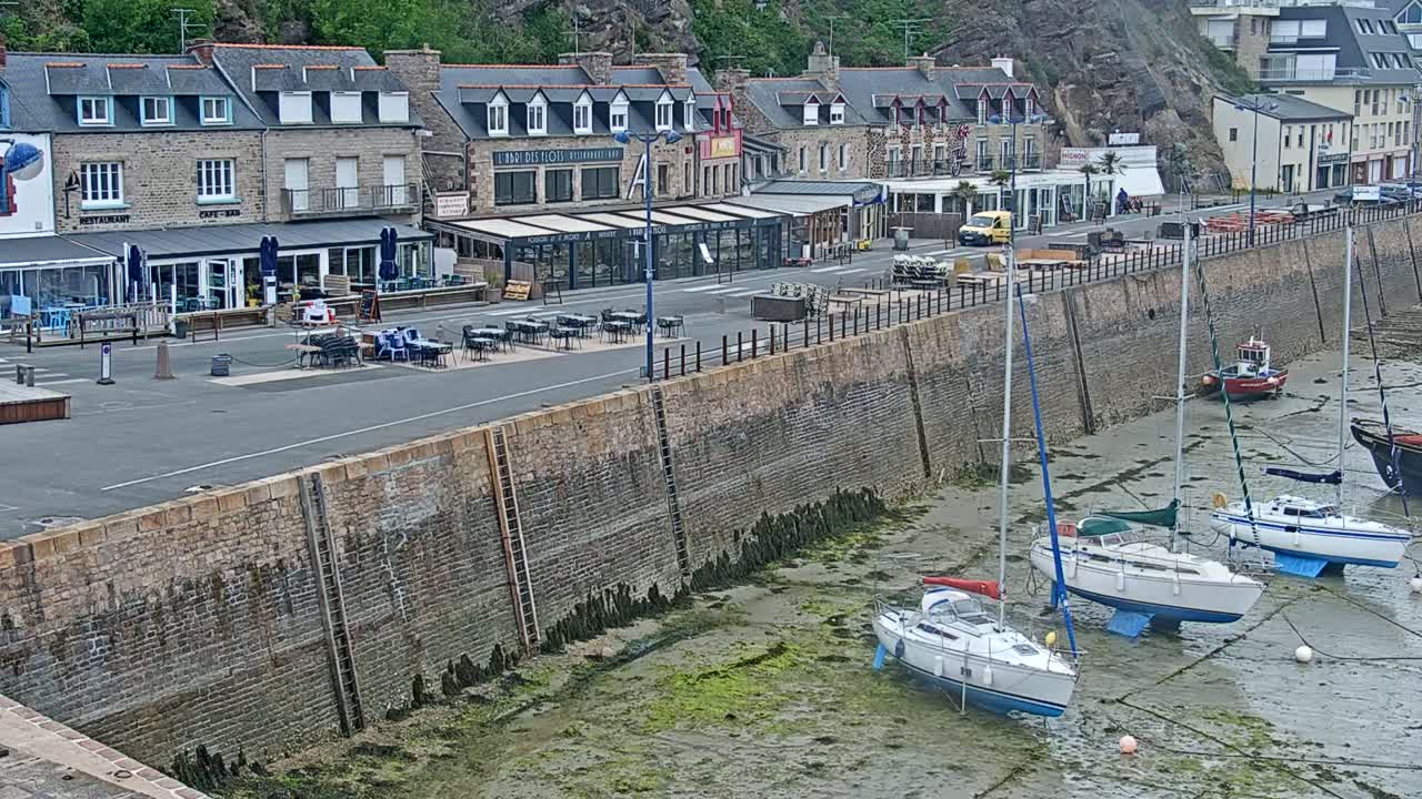 A stone harbor wall with several sailboats resting on the mud flats at low tide, in front of a row of seaside buildings and a paved area with outdoor seating.