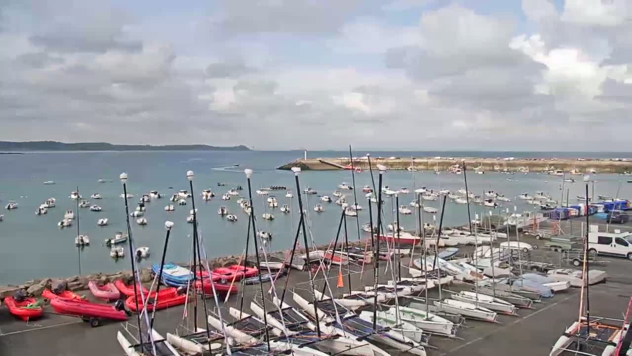 Numerous sailboats and catamarans are docked in a harbor on an overcast day, with additional boats anchored in the calm water beyond.