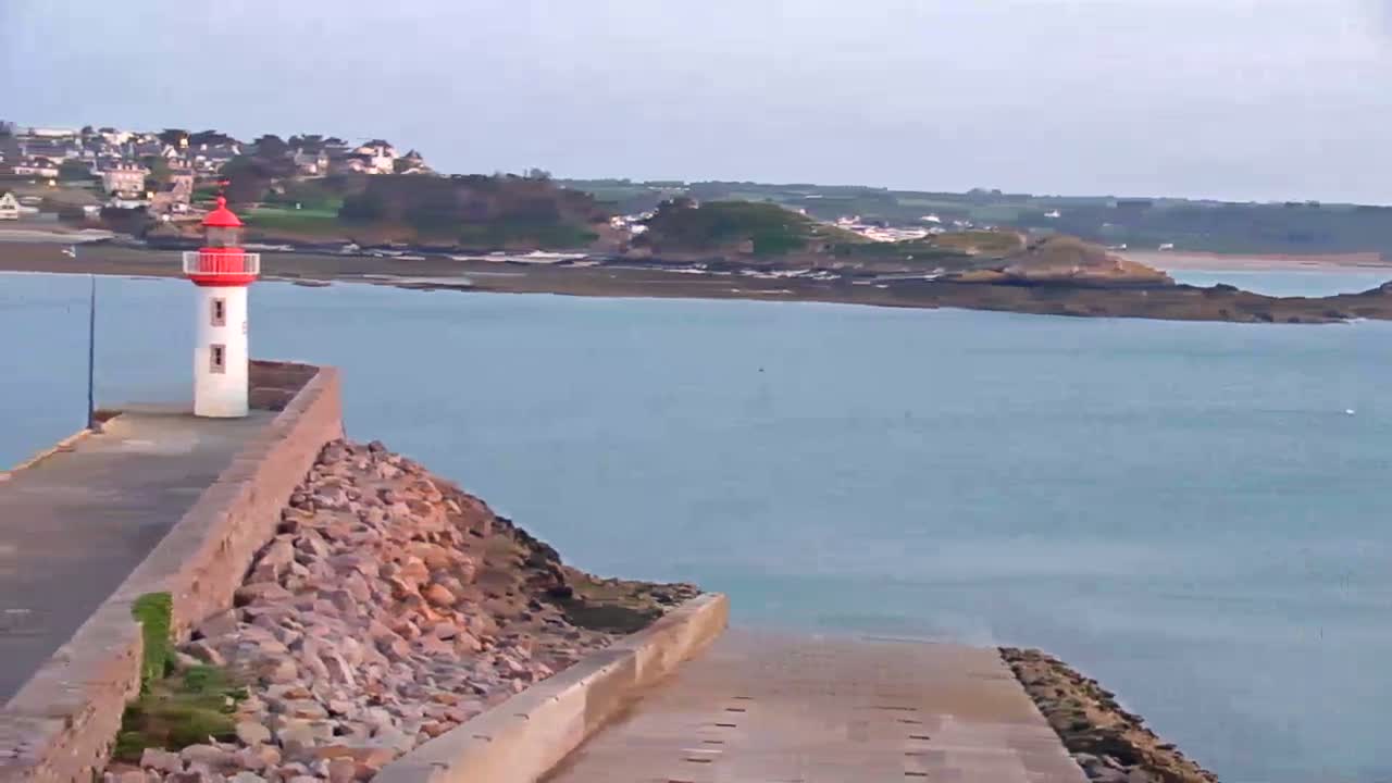 A red and white lighthouse stands on a stone jetty overlooking a calm body of water and a distant coastline under a clear sky.
