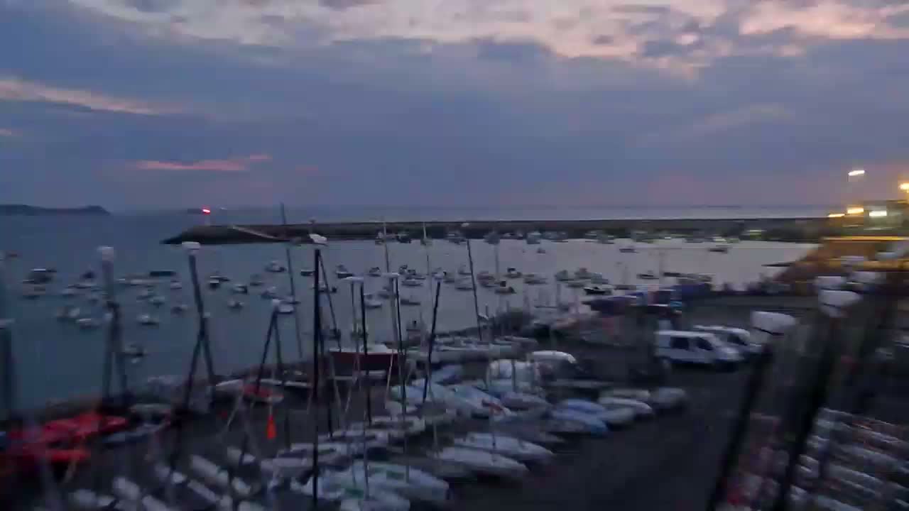 Many sailboats are docked in a harbor at dusk under a cloudy sky.