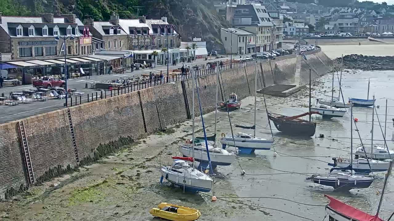 A seaside town with buildings lining a waterfront, several sailboats are docked at low tide on a muddy shore.