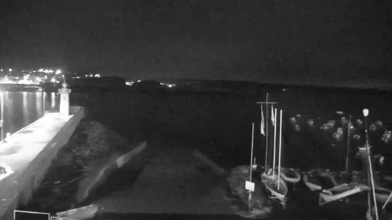 A nighttime view of a harbor with several small boats moored near a stone pier and a small lighthouse, under a clear, dark sky.
