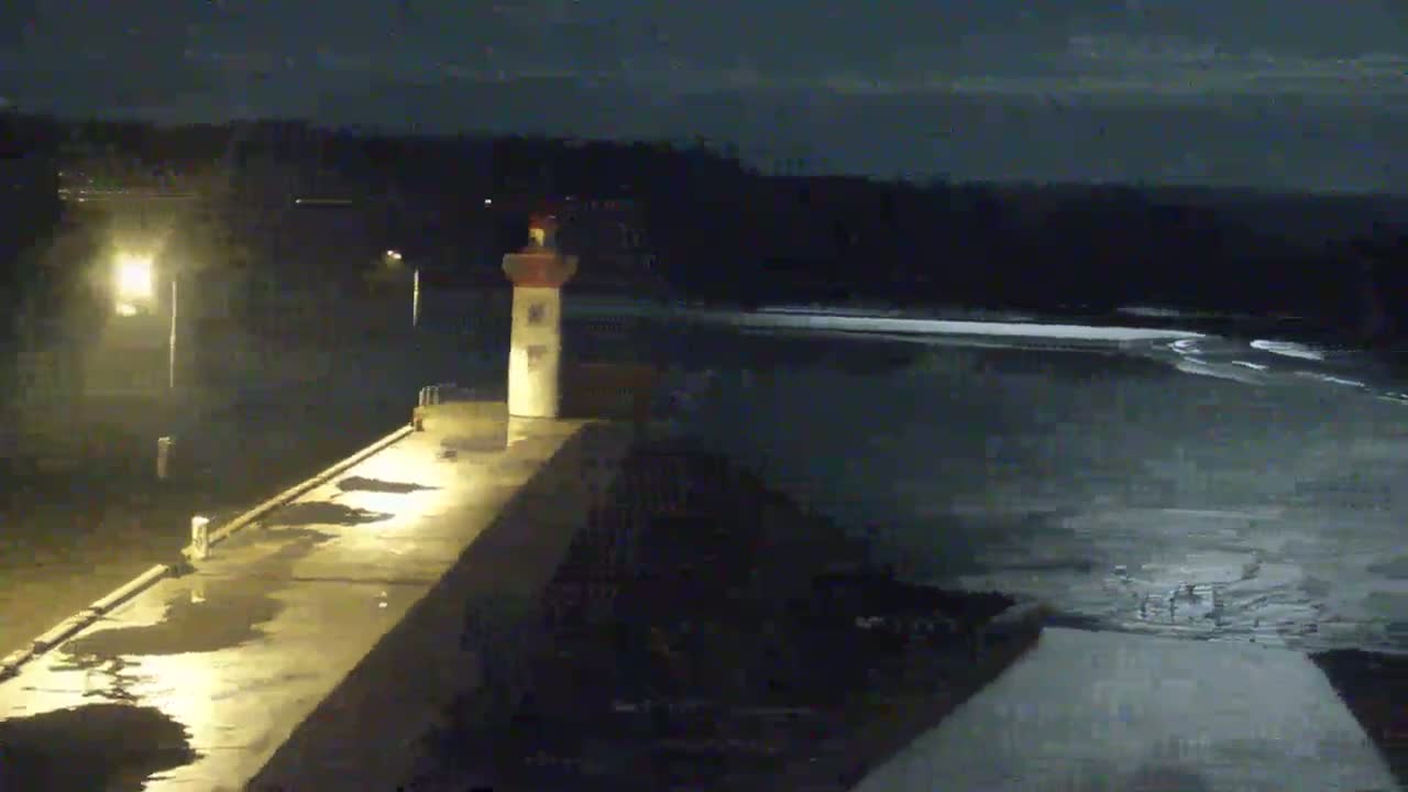 A small lighthouse sits on a wet pier at night, overlooking dark, choppy water under a cloudy sky.