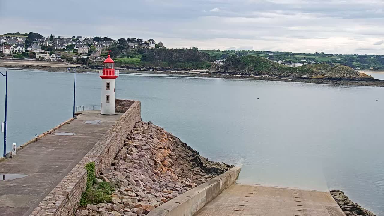 A red and white lighthouse stands on a stone jetty overlooking a calm body of water under a cloudy sky, with a small town and island visible in the distance.