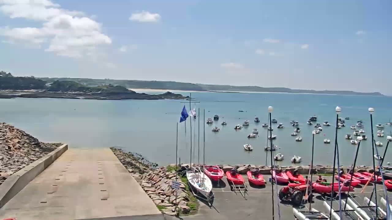 A calm, sunny day overlooks a harbor filled with numerous boats, a boat ramp, and several red sailboats parked on land.