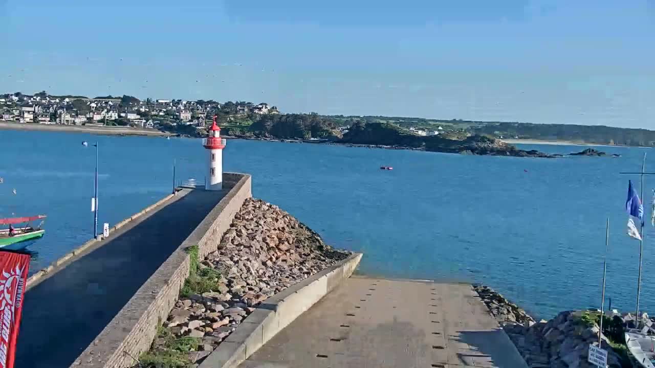 A red and white lighthouse stands on a stone jetty overlooking a calm blue ocean, with a town visible in the distance under a clear sky.