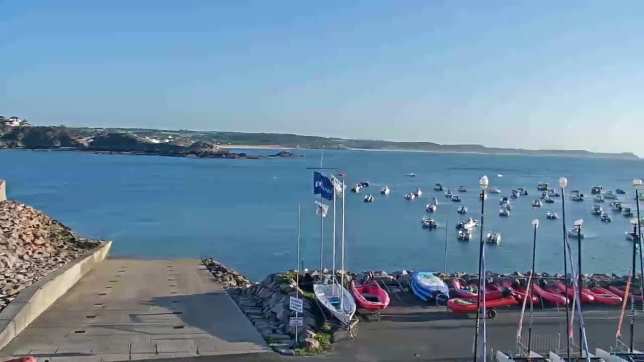 A calm, sunny day shows a harbor filled with numerous boats, some moored and others at anchor in the water, with a boat ramp and additional vessels beached nearby.