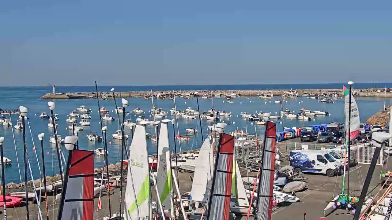 A harbor filled with numerous sailboats and motorboats under a clear blue sky, with additional sailboats and some vehicles parked on the land.