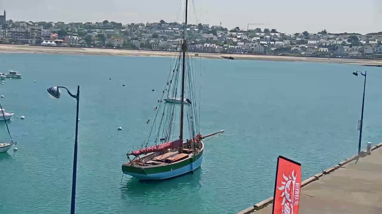 A sailboat is moored in calm, teal water near a seaside town on a sunny day.