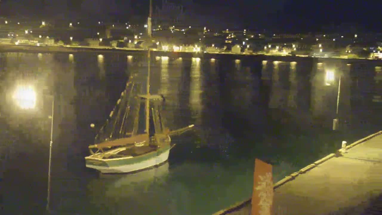 A sailboat is moored in calm water at night near a town's illuminated waterfront.