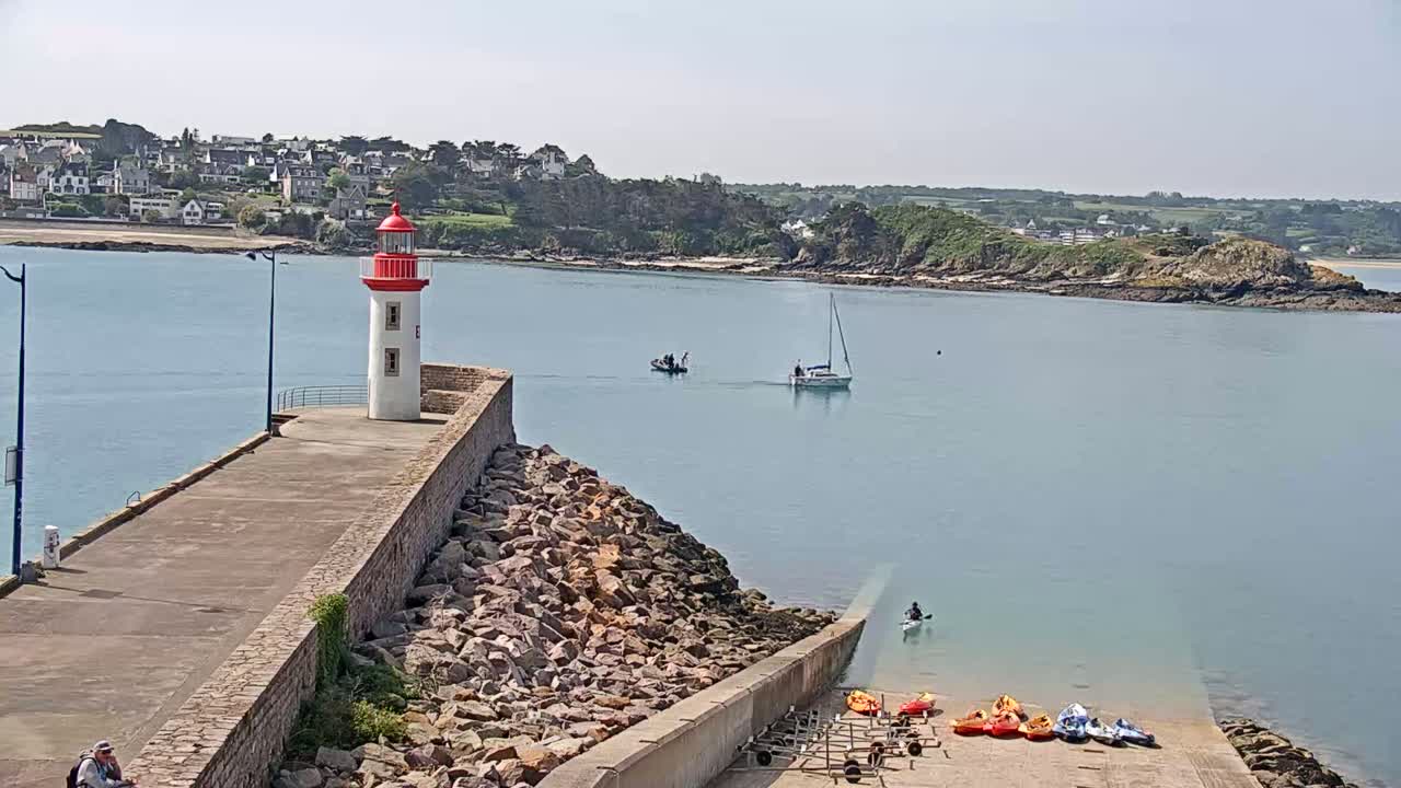 A red and white lighthouse stands on a stone jetty overlooking a calm body of water with a sailboat and kayakers, a small town visible in the distance on a sunny day.