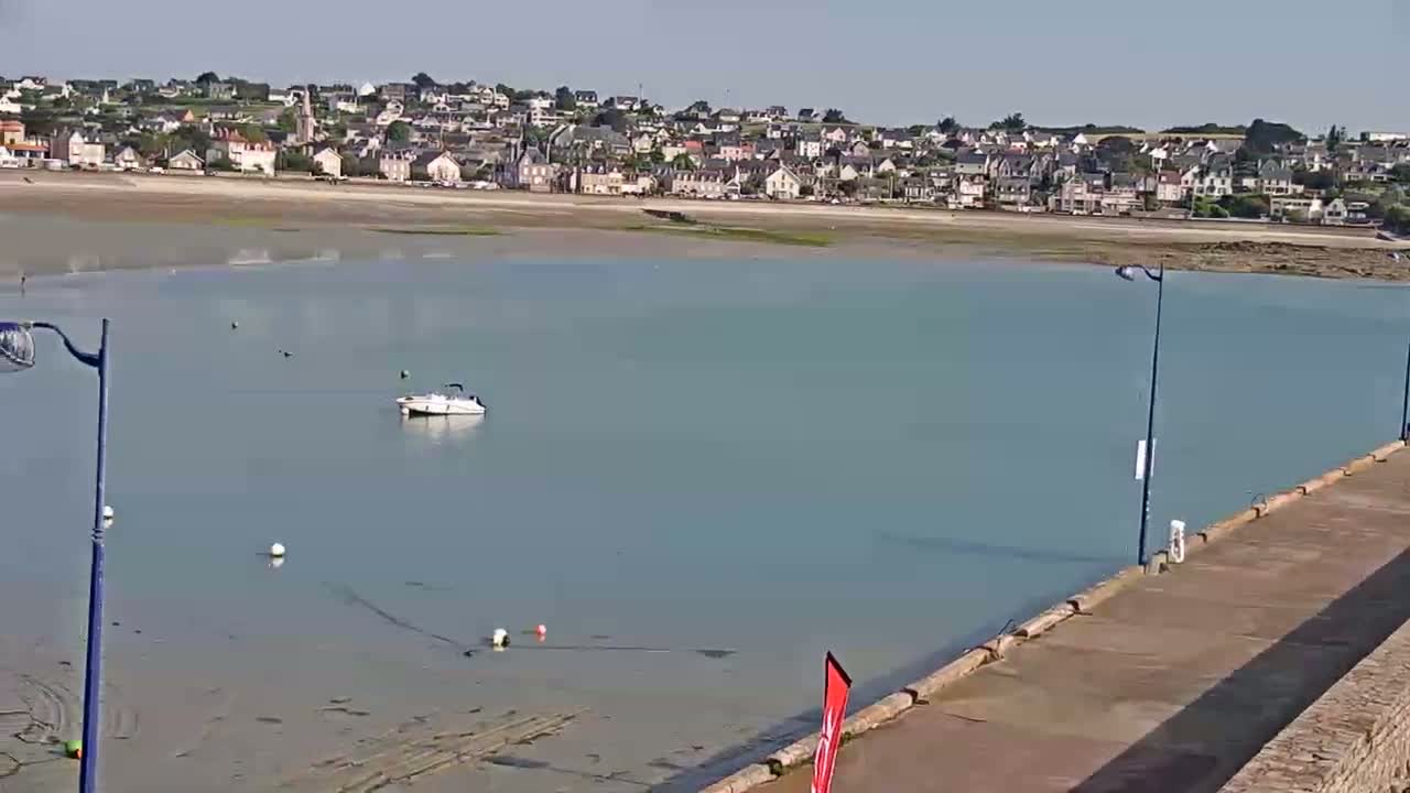 A small motorboat is anchored in calm, light-blue water near a seaside town on a sunny day.