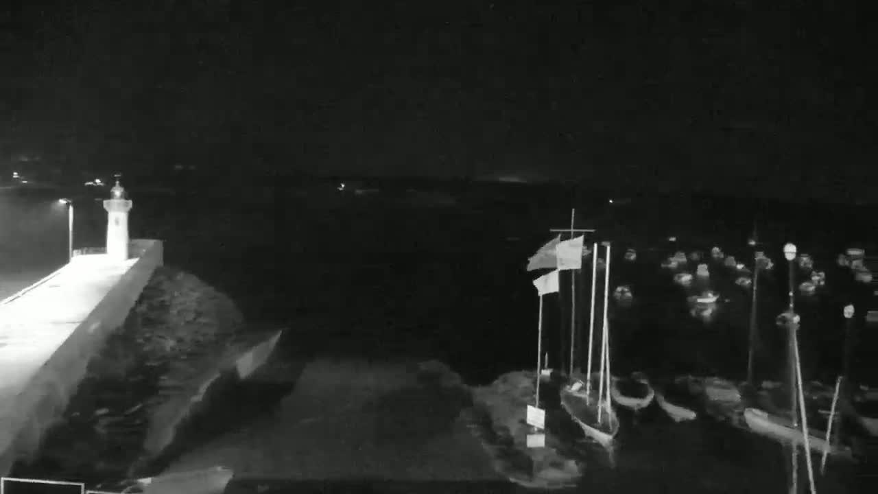 A nighttime, low-light view shows a harbor with numerous boats moored, a small lighthouse on a stone pier, and calm water.