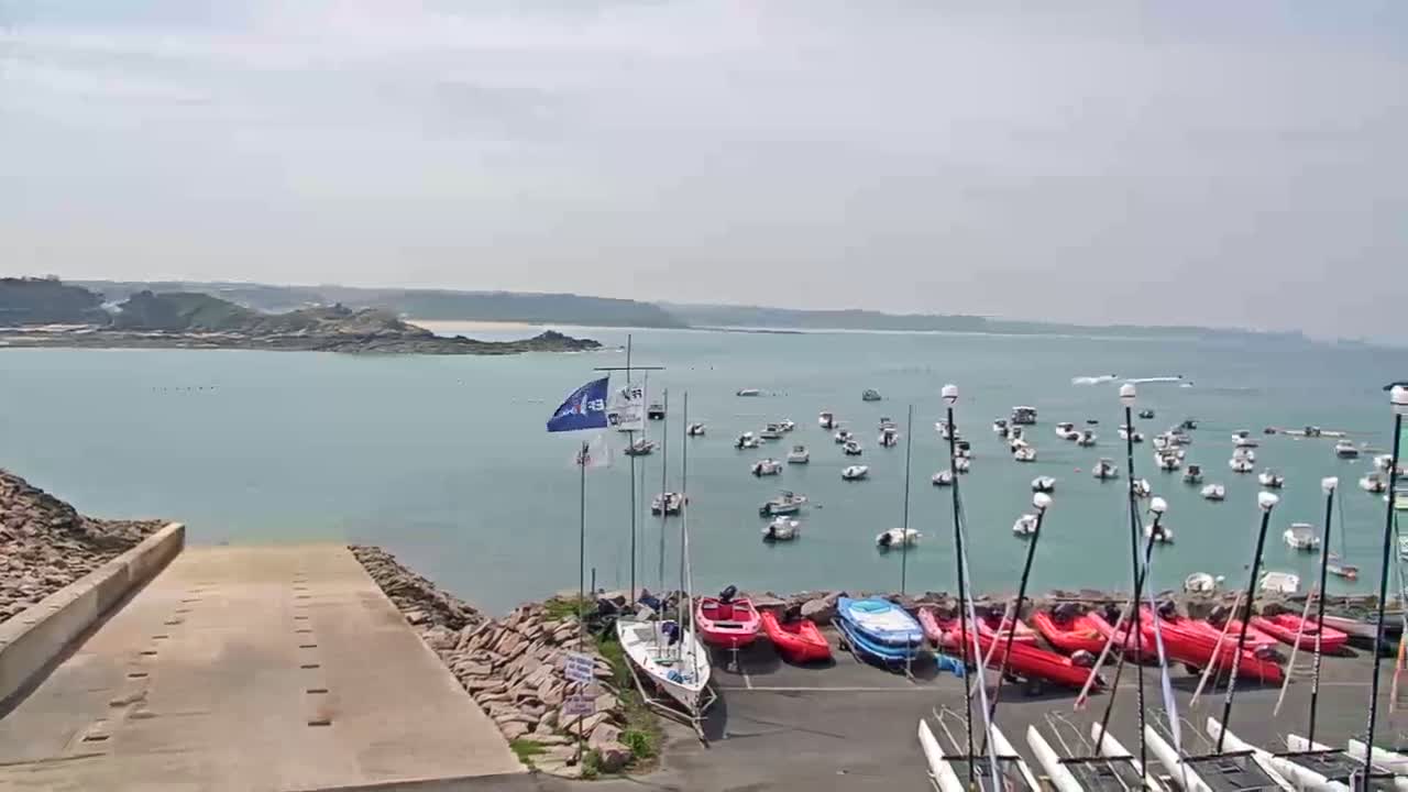 A calm, light-blue body of water dotted with numerous small boats and sailboats is viewed from a rocky shoreline, where additional boats and catamarans are parked.