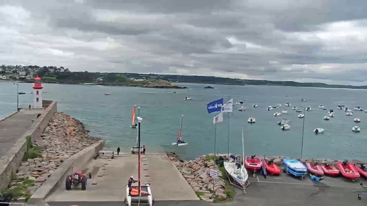 A harbor scene under an overcast sky shows a lighthouse on a rocky jetty, numerous sailboats and small motorboats in the water, and several small boats and a tractor parked on land.