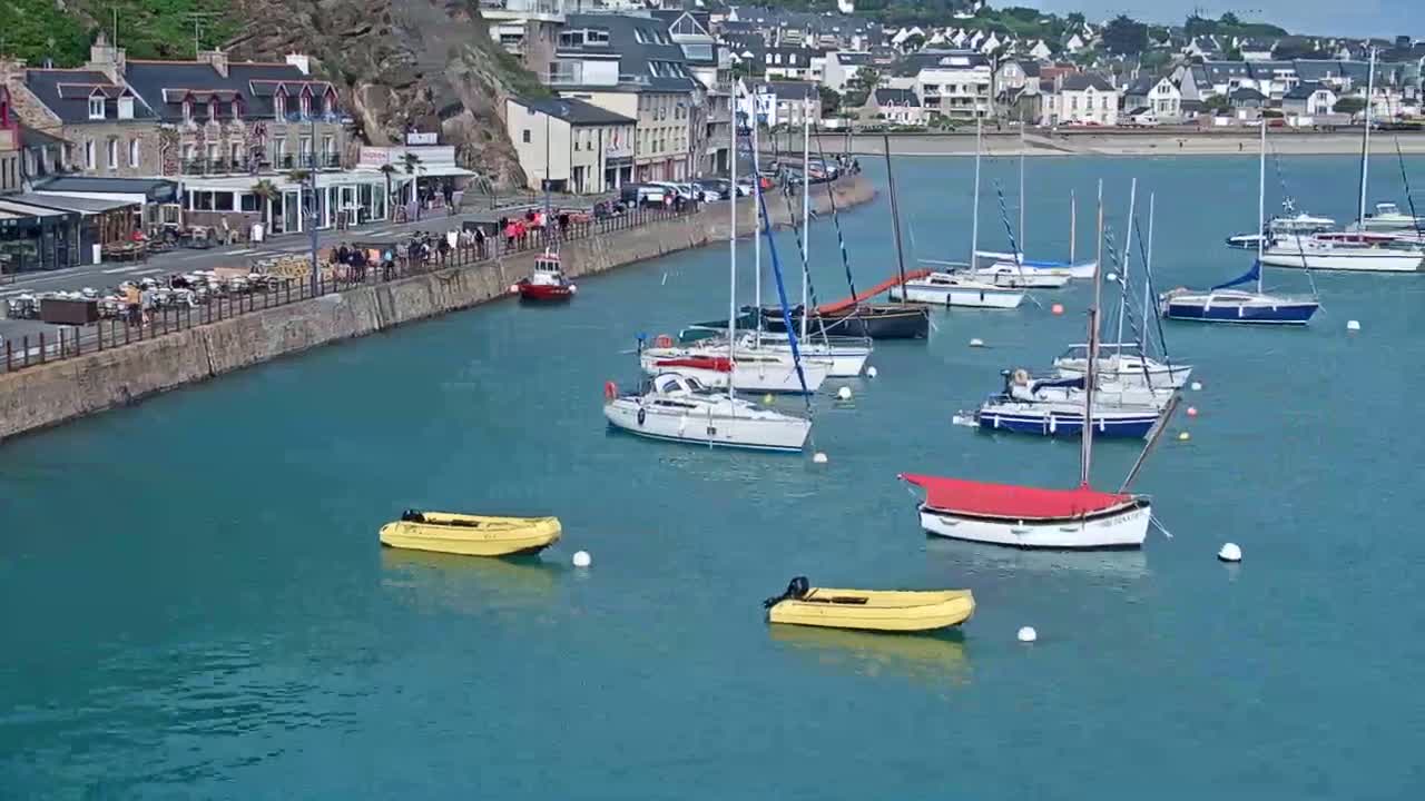 Numerous sailboats and small motorboats are moored in a calm, teal harbor next to a waterfront town with buildings and people visible along the shore.