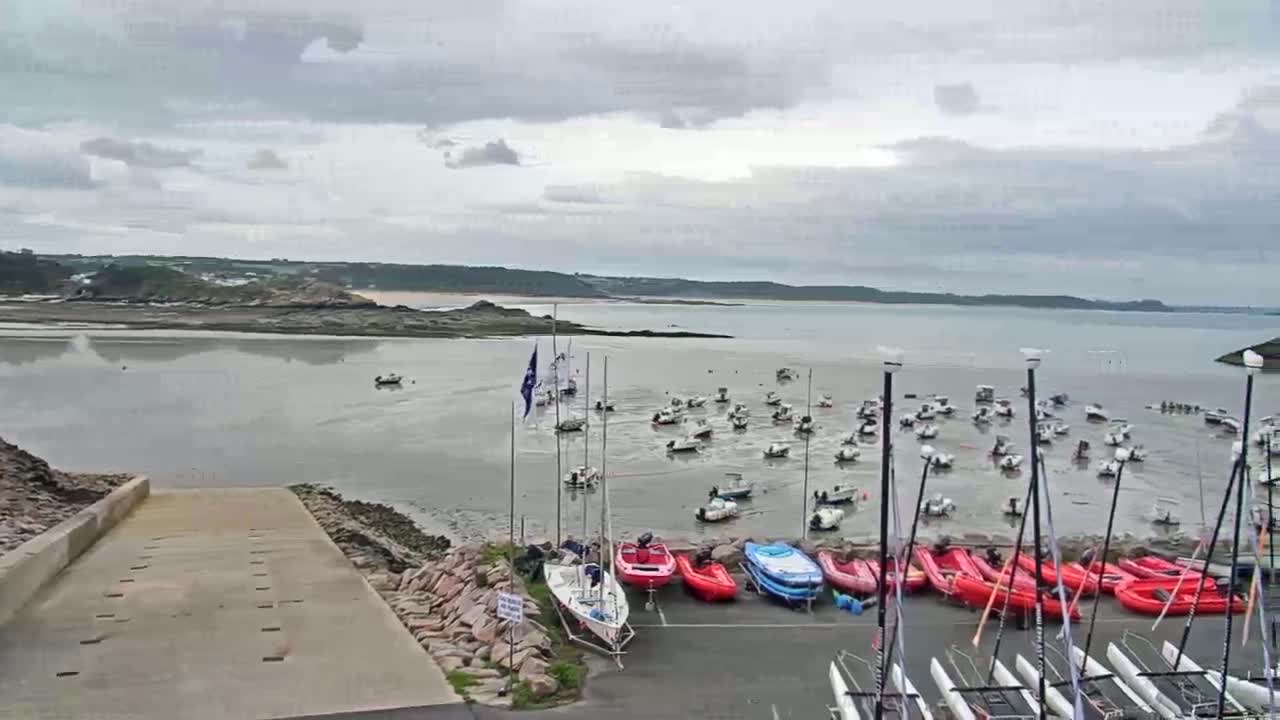 A harbor filled with numerous sailboats and motorboats sits at low tide under a cloudy sky, with a calm body of water and distant land visible in the background.
