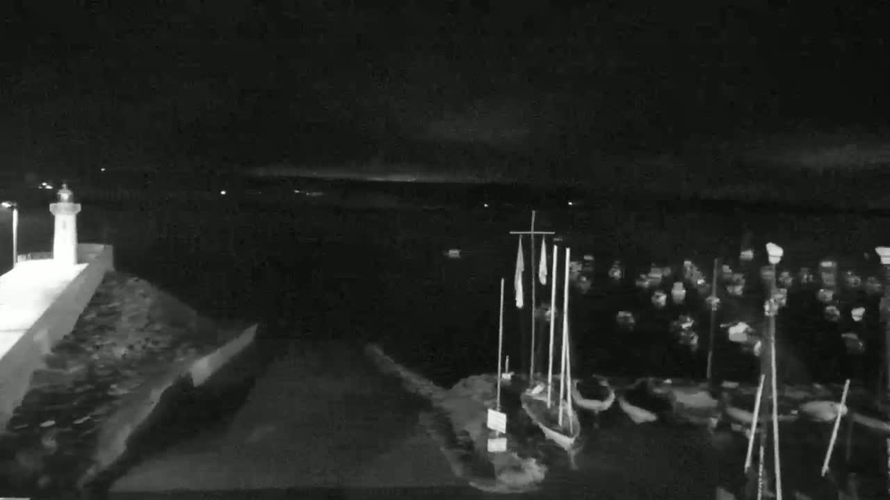 A nighttime, low-light view shows a small lighthouse on a stone jetty, with numerous boats moored in the dark water beyond.