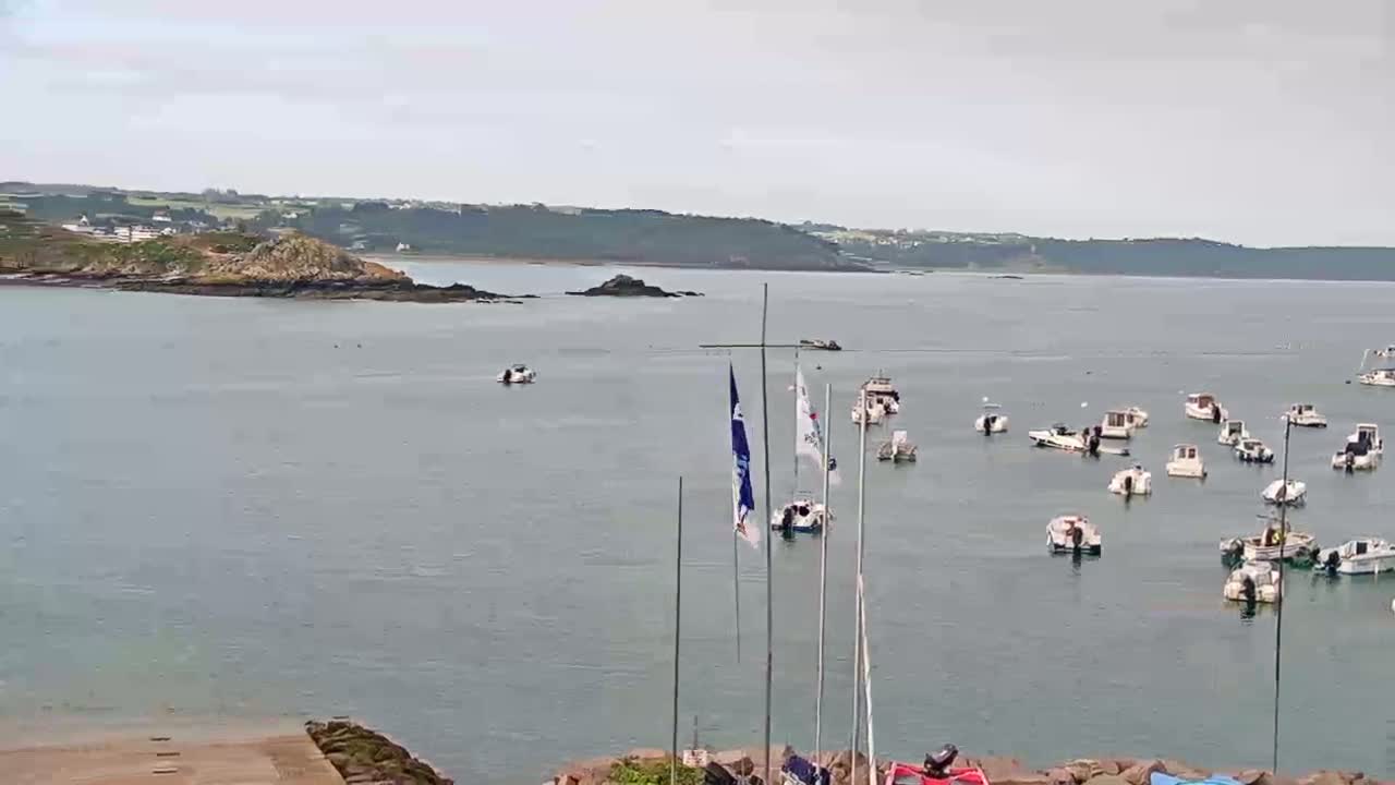A calm bay with numerous small boats moored, a rocky islet in the near distance, and a hazy sky.