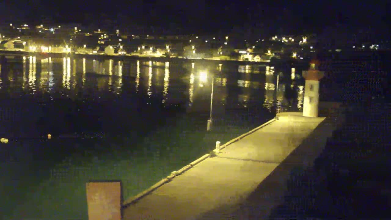 A nighttime view of a pier extending into a calm body of water reflecting the lights of a town across the water, with a small lighthouse at the pier's end.